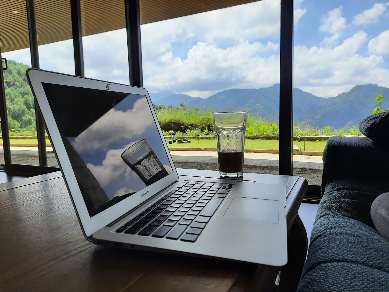 A laptop open on a table by a window overlooking hills, a coffee within reach.